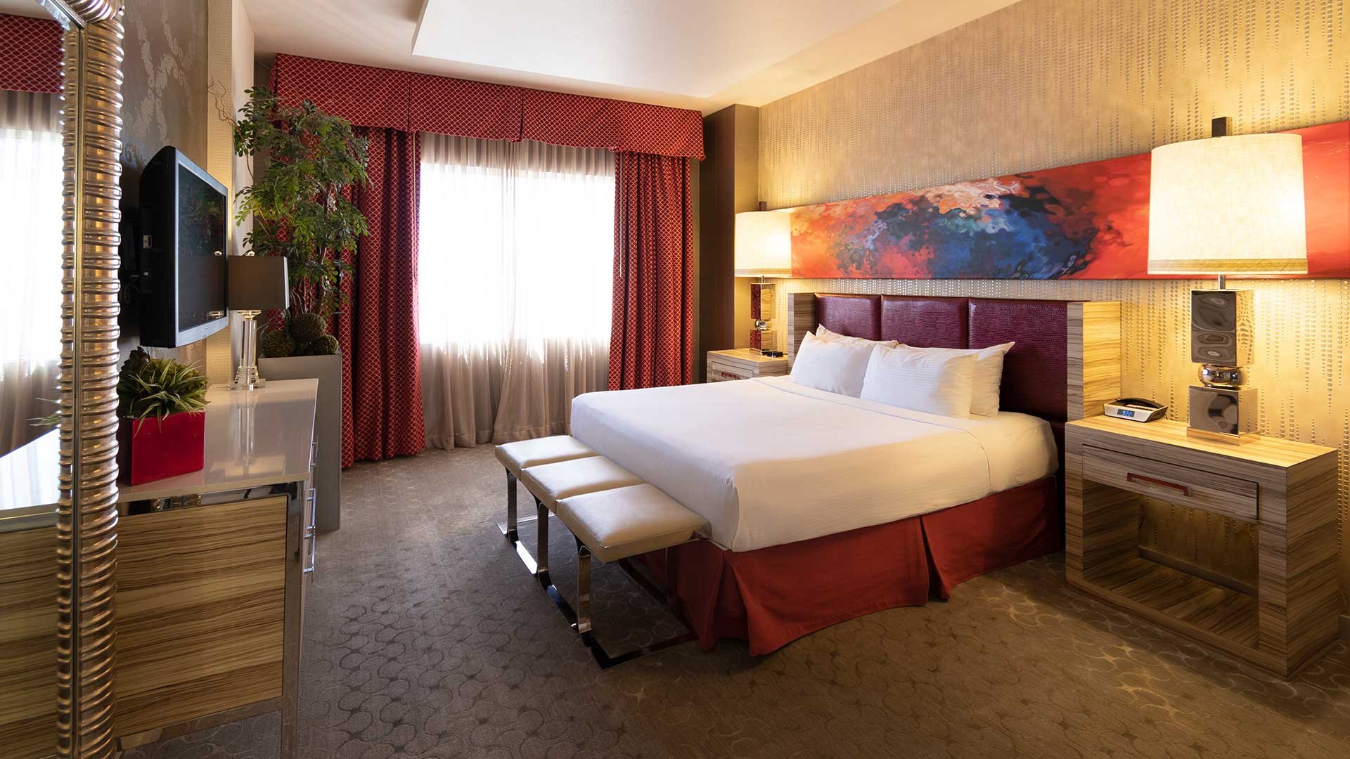 A hotel room with a king-sized bed, bench, TV, plants, and artwork