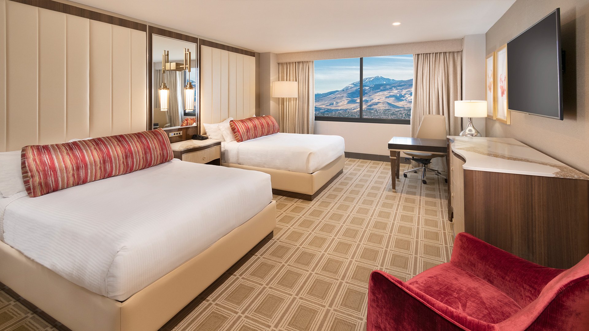 A hotel room with two beds, a desk, a TV, and a chair, with a view of mountains outside the window.
