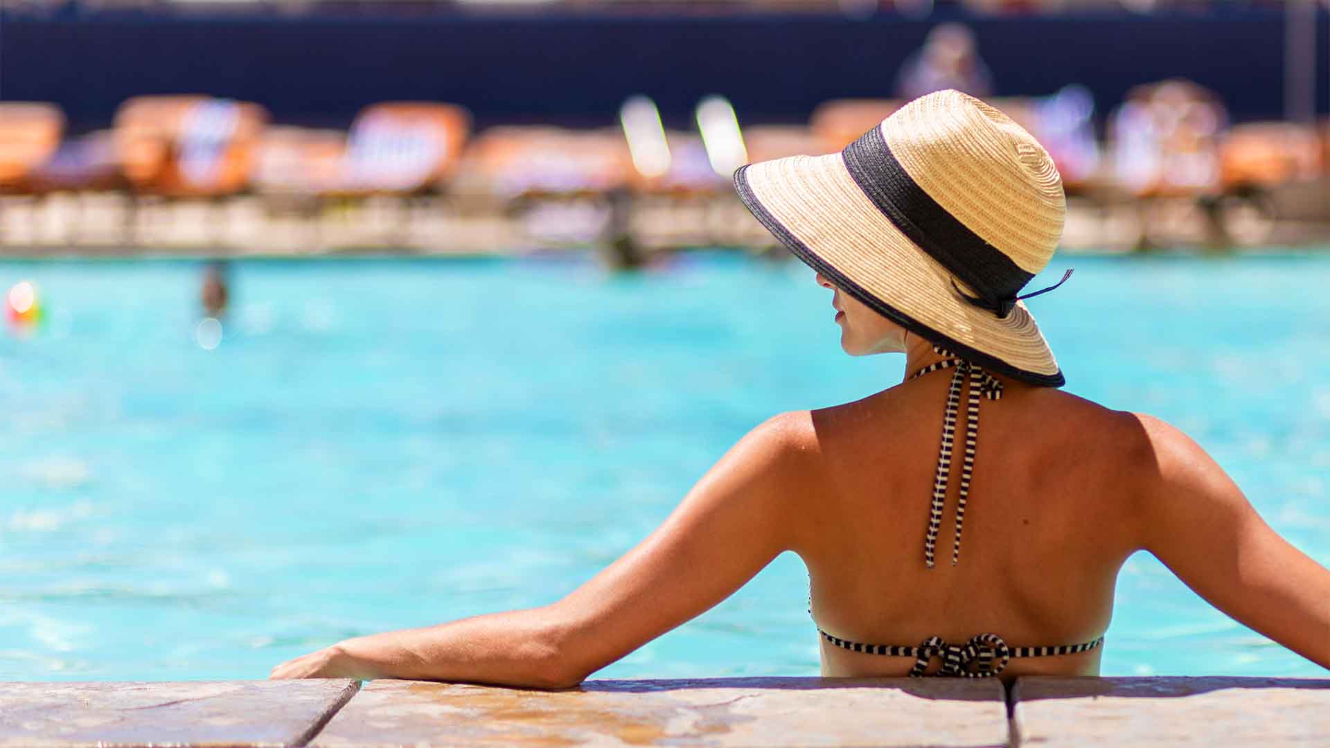 A woman in a hat relaxes at the edge of an outdoor pool.