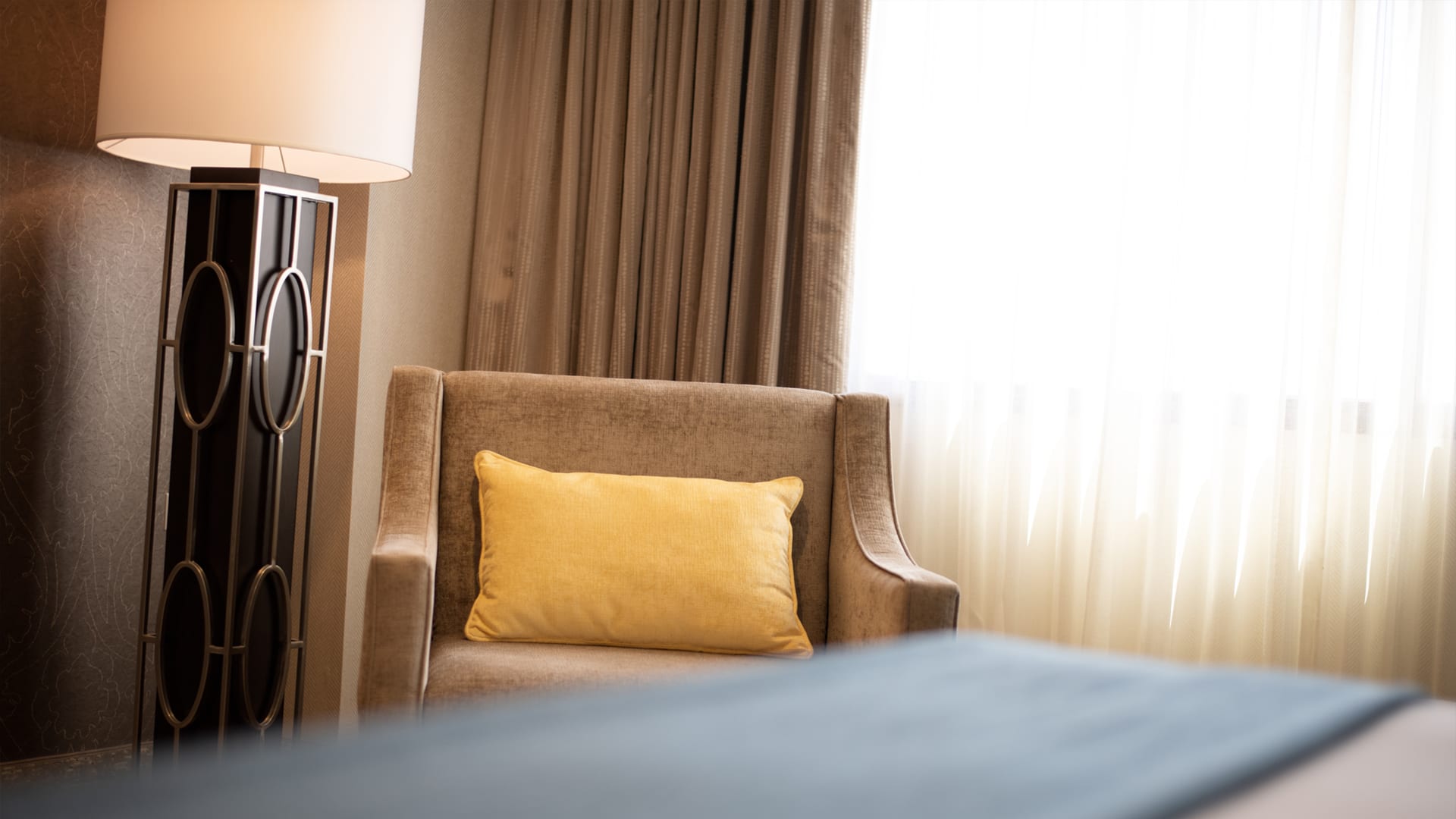 A chair and lamp in a hotel room with a bed and window