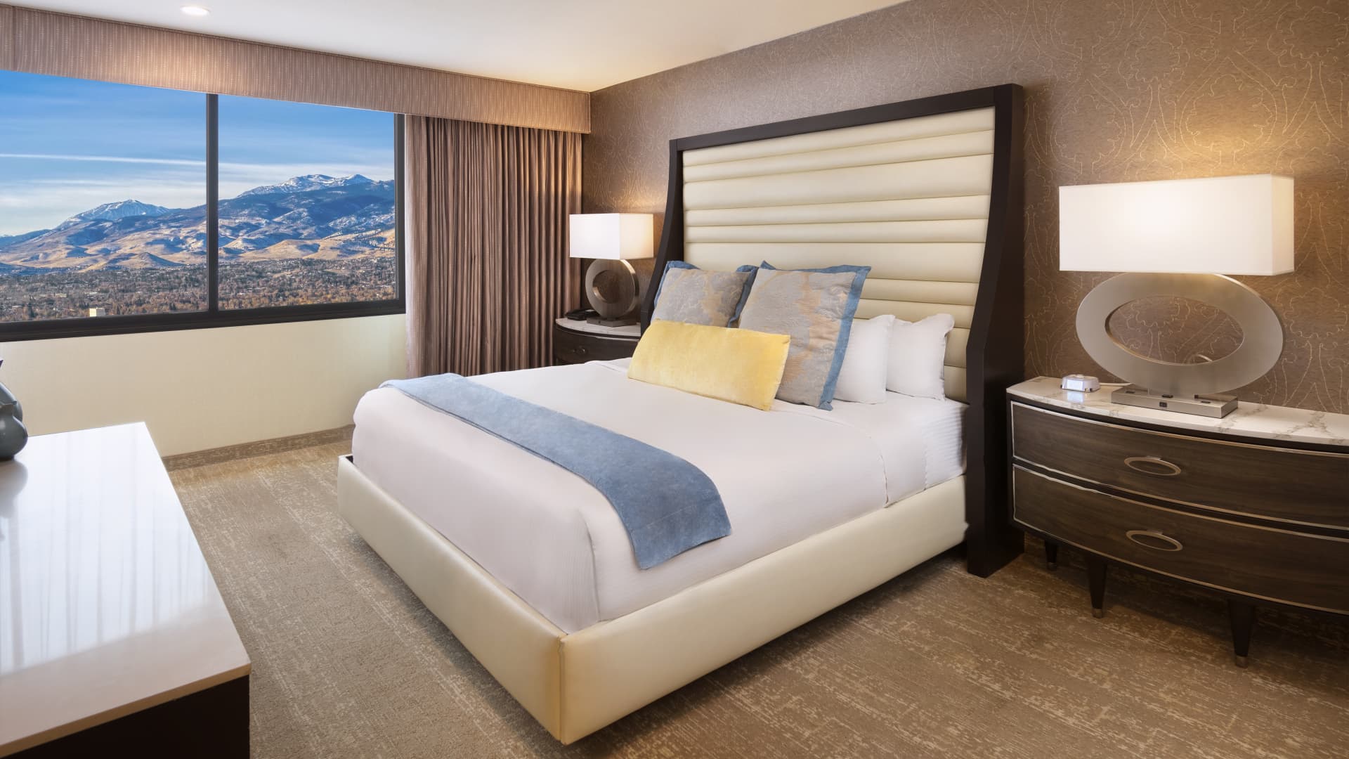 A hotel room with a large bed, nightstand, and window showing a mountain view.