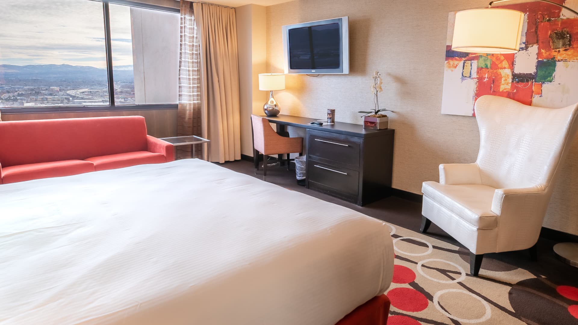 A hotel room with a king-sized bed, red sofa, desk, chair, and TV on the wall.