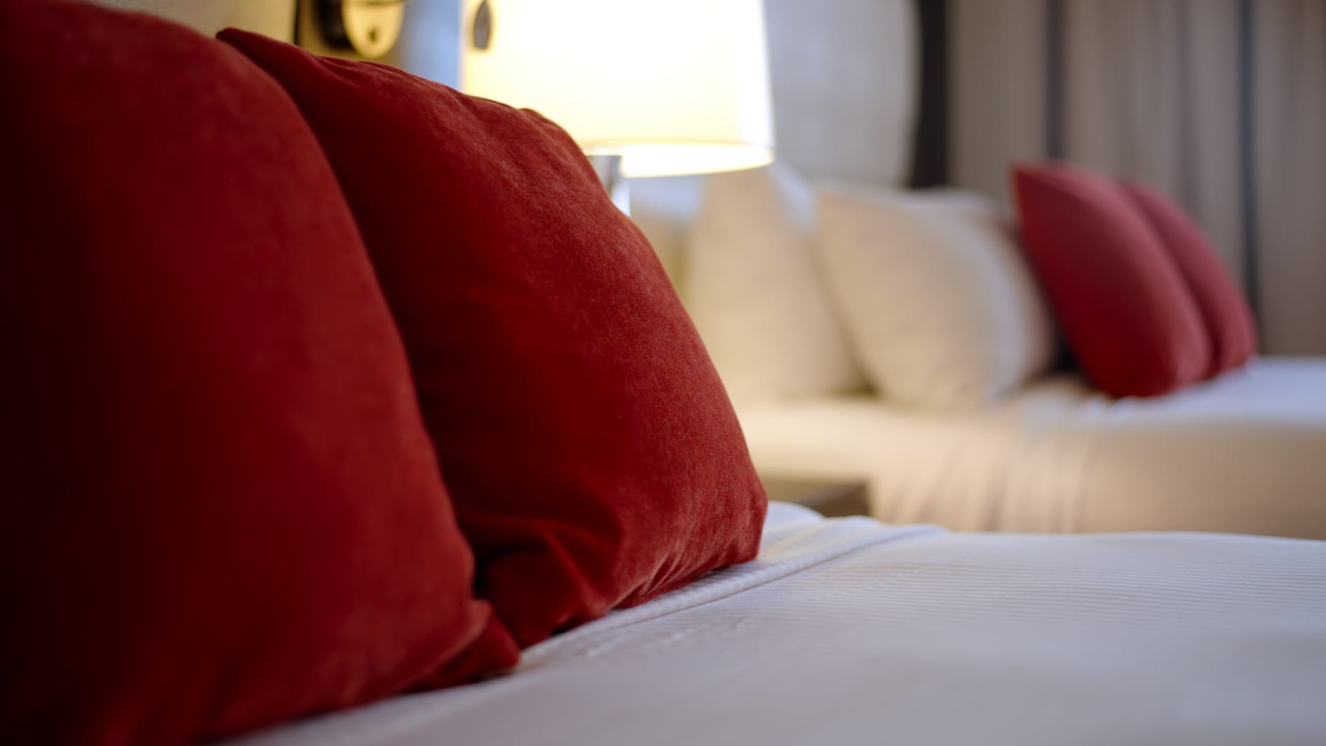 A close-up of red pillows on a bed with white bedding and a lamp in the background.