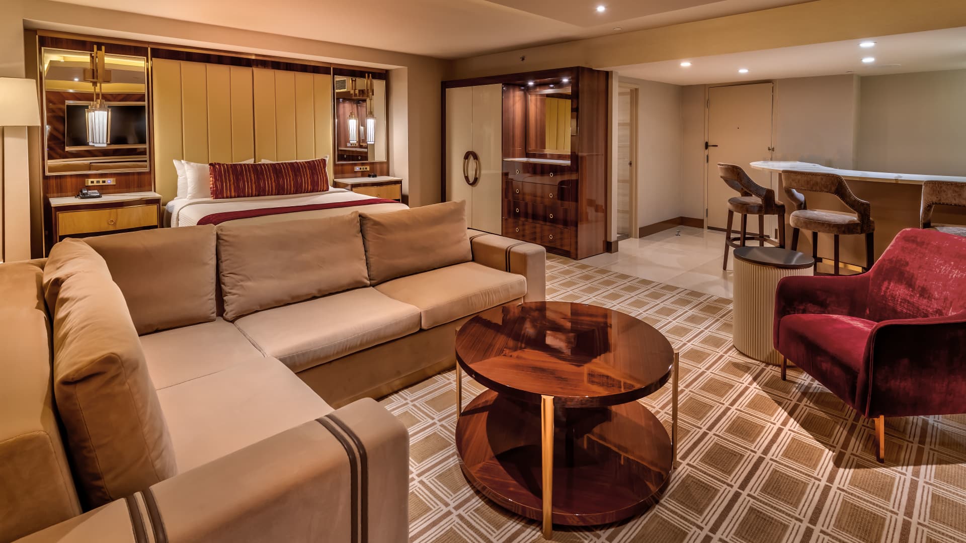 A wide view of a hotel suite's living area with a sofa, coffee table, armchair, bar, and bed in the background.