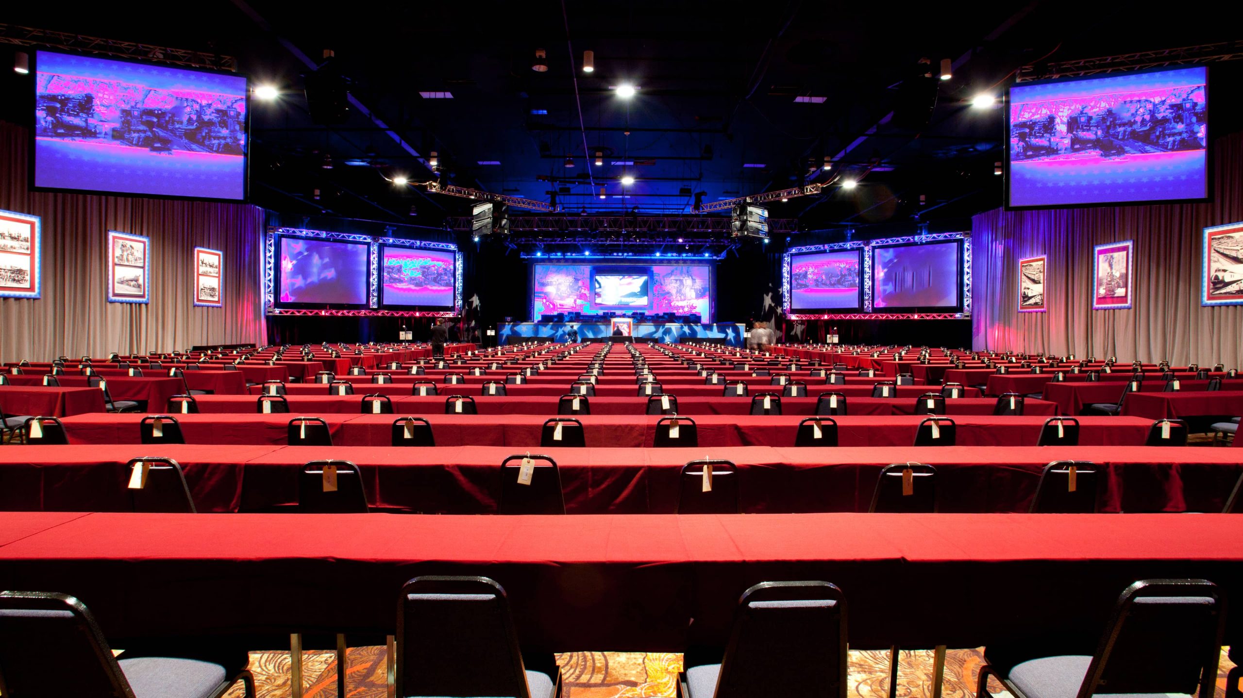 A large meeting room with red tables, black chairs, and large screens on the walls.