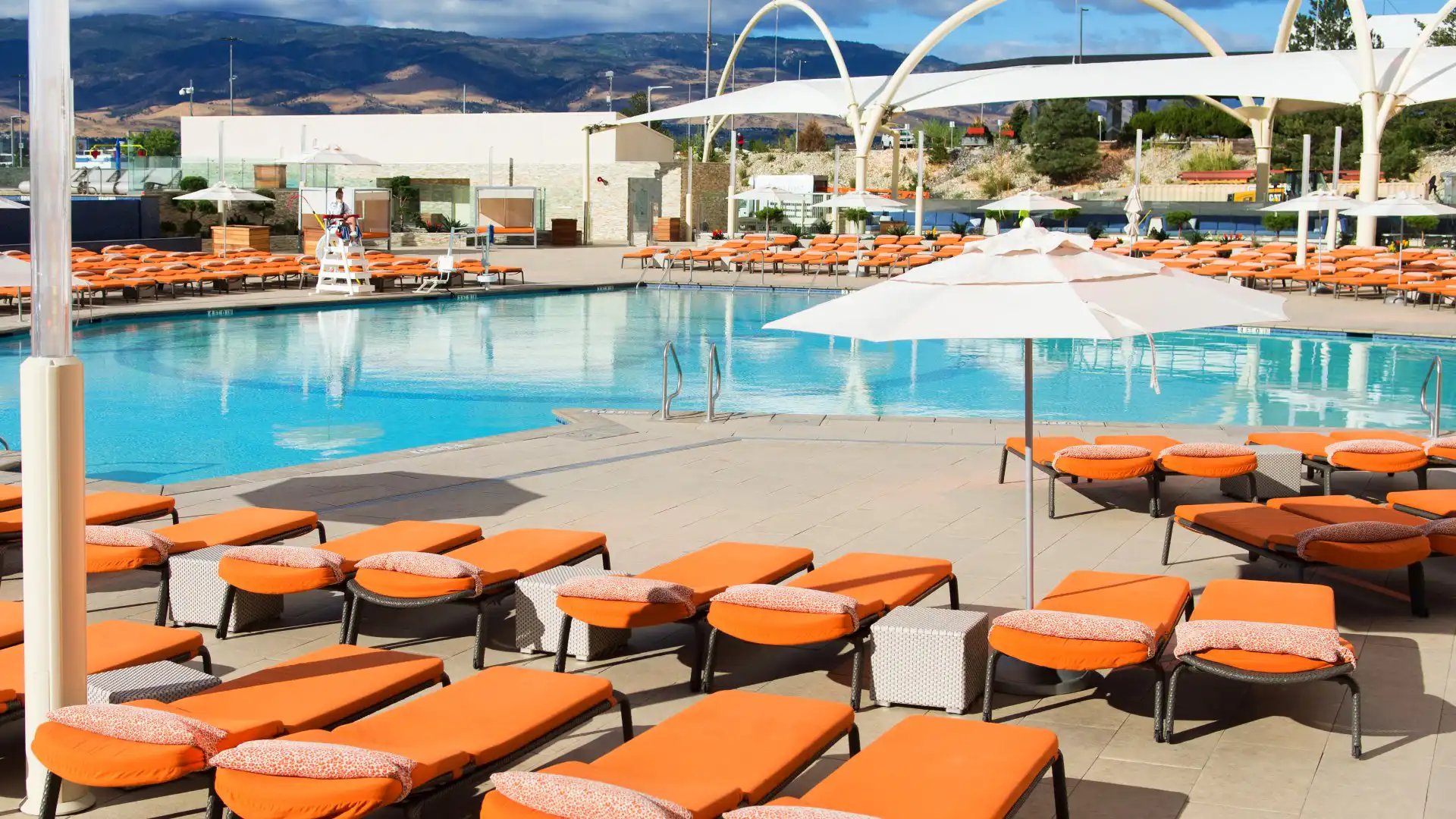 A poolside area with orange lounge chairs, white umbrellas, and a mountain view.